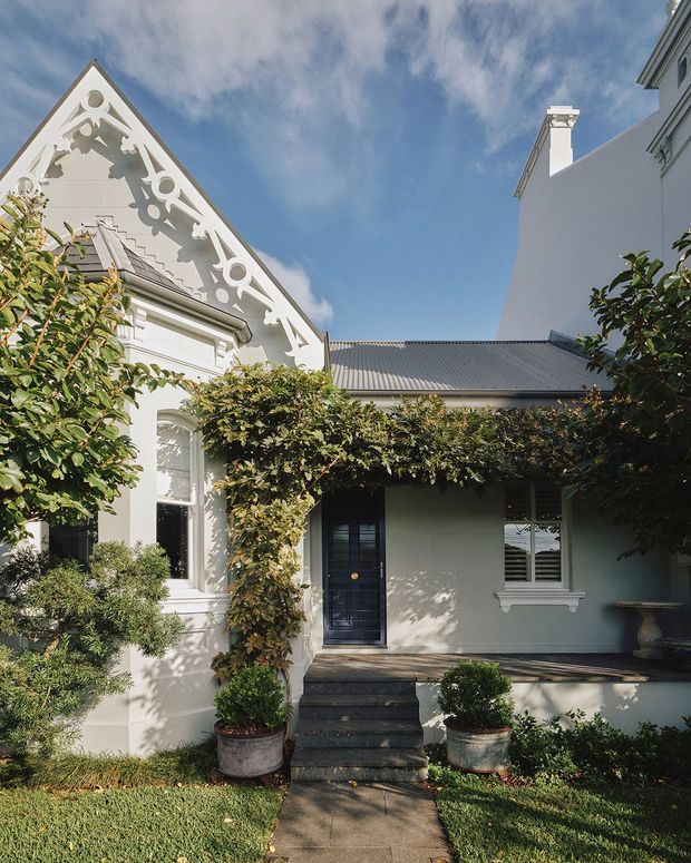Exterior view of a refined heritage-style residence with a white rendered facade, ornate decorative gable detailing, arched window surrounds, and a dark corrugated roof. A central entry porch is framed by climbing greenery, with steps, potted plants, and a paved path leading through the landscaped garden.