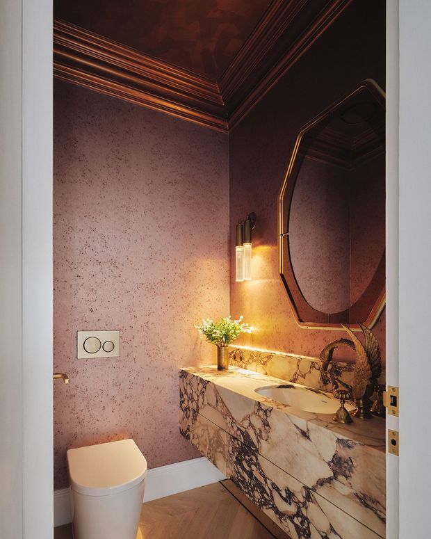 A compact, luxurious powder room viewed through an open doorway. The space features textured mauve-pink wall finish, an ornate dark glossy ceiling with layered trim, a dramatic veined marble floating vanity with integrated basin, sculptural black and brass tapware, a large angular framed mirror, warm wall lighting, and a contemporary white toilet. Timber flooring and brass-accented fittings complete the moody, opulent interior.