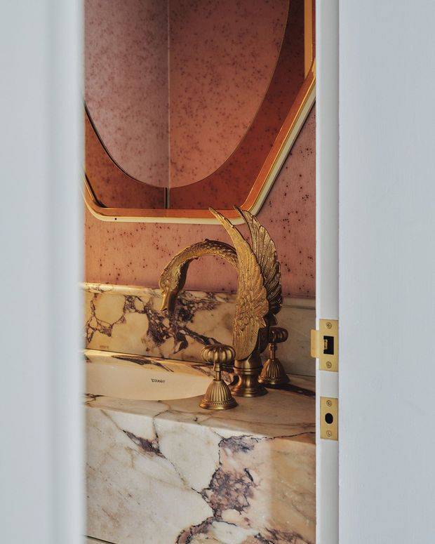 Close view through a partially open doorway into an ornate bathroom. A sculptural antique-style brass faucet with shell-like detailing sits on a richly veined marble vanity, beneath a curved copper-toned mirror or wall panel. The composition is luxurious and highly decorative, with warm metallic accents and dramatic natural stone.