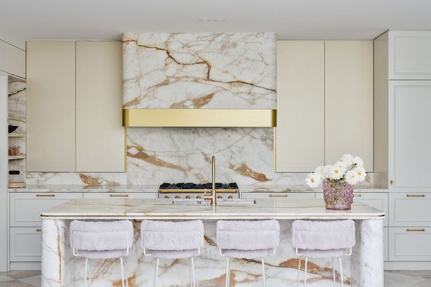 Refined contemporary kitchen featuring a large waterfall island clad in dramatic white marble with warm gold veining, matching marble splashback and rangehood surround, pale beige built-in cabinetry, and brushed brass accents. Four upholstered bar stools and a floral arrangement sit at the island, creating a light, luxurious entertaining space.