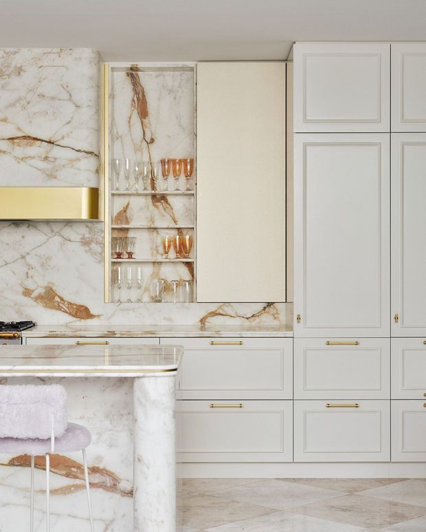 A refined, light-toned kitchen with extensive veined marble on the island, splashback and wall surfaces. Pale grey-white shaker-style cabinetry with brass pulls runs across the wall, alongside a glass-front drinks cabinet displaying stemware. The island has a rounded marble-clad end and a pale upholstered stool. Geometric pale stone flooring and warm brass details create an elegant, luxurious finish.