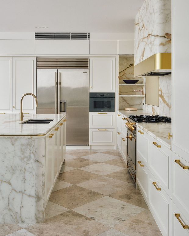 A refined galley-style kitchen with white shaker cabinetry, brass hardware and fixtures, a large waterfall island clad in heavily veined white marble, stainless-steel appliances, and a matching marble splashback and rangehood surround. The floor features large diagonal checkerboard tiles in warm beige and cream tones. Open shelving, integrated refrigeration, a gas cooktop, and built-in oven complete the bright, luxurious space.