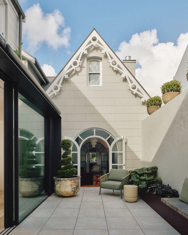 A bright, enclosed courtyard or terrace framed by a white gabled building with ornate decorative bargeboard and arched windows and doors. Large pale tiled paving, dark-framed glazing, sculpted topiary, potted plants, a green lounge chair and a built-in planter bench create a refined heritage-inspired outdoor seating area. The image is photographed outdoors under a blue sky.
