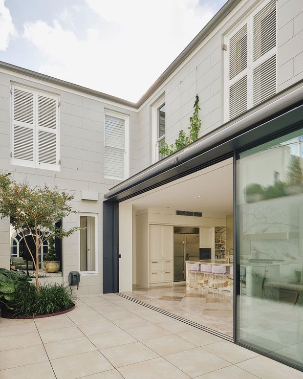 Bright courtyard-facing view of a refined two-storey residence with a pale rendered or panelled facade, louvred window shutters, and a large dark-framed glazed opening connecting the indoor kitchen to the tiled outdoor terrace. The kitchen features light cabinetry, a stone island, and marble-look finishes. Landscaping is limited to a planted courtyard bed beside the paved terrace.