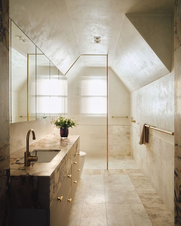 A refined bathroom with a marble-clad vanity, integrated basin and brushed brass tapware, large mirrored wall, glass shower enclosure with brass framing, and pale stone wall and floor tiles. The room features a dramatic sloped ceiling, skylight-like recessed lighting, fluted translucent windows, and warm neutral finishes.