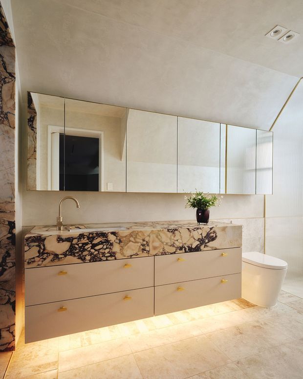 A refined contemporary bathroom featuring a wall-mounted double vanity with pale cabinetry, small brass-gold pulls, and a dramatic veined natural-stone benchtop and side panel. A wide mirrored cabinet spans the wall above the vanity, with a sculptural brushed brass tap, a white wall-hung toilet, large pale floor tiles, and warm concealed under-vanity lighting. The palette is warm beige, cream, brown, black, and brass, with a small plant adding greenery.