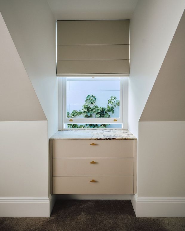 A compact built-in dressing or storage nook beneath a sloped roof, featuring three beige drawers with small brass-gold pulls, a pale marble-look countertop, white sloping walls, and a centered window with a beige Roman blind. Dark carpet covers the floor, and greenery is visible outside the window.