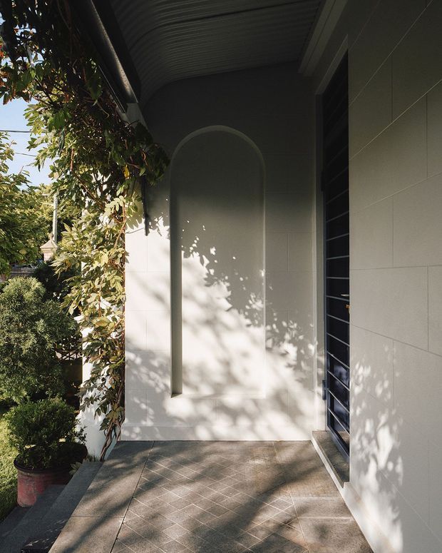 A shaded residential entrance porch with a tiled floor, pale rendered or panelled walls, a tall arched recessed feature, and a dark framed louvre-style door. Dense planting and climbing greenery border the walkway, casting dappled shadows across the facade and paving.