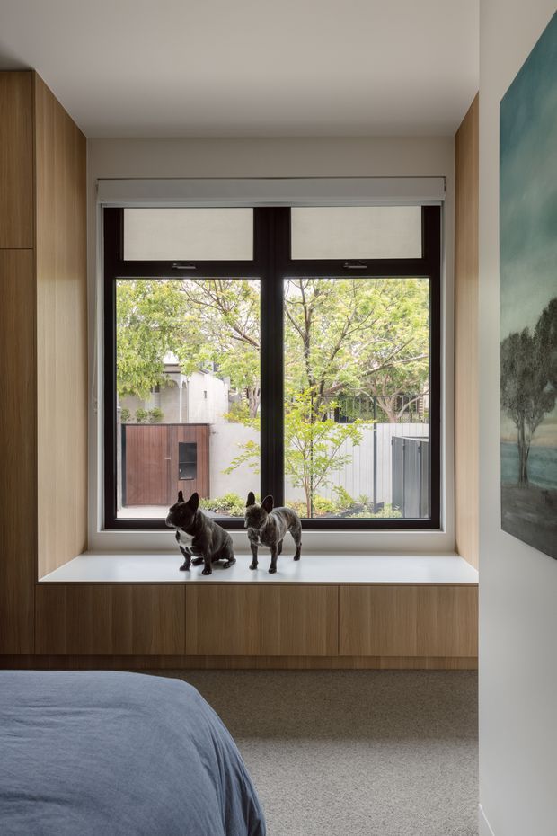 A calm contemporary bedroom corner featuring a large dark-framed window with roller blinds, a built-in timber window seat and surrounding full-height timber cabinetry. A bed with blue-grey bedding is partially visible in the foreground, while sculptural dog figures sit on the window seat. The window overlooks a leafy urban courtyard or garden, and a large landscape artwork occupies the adjacent wall.