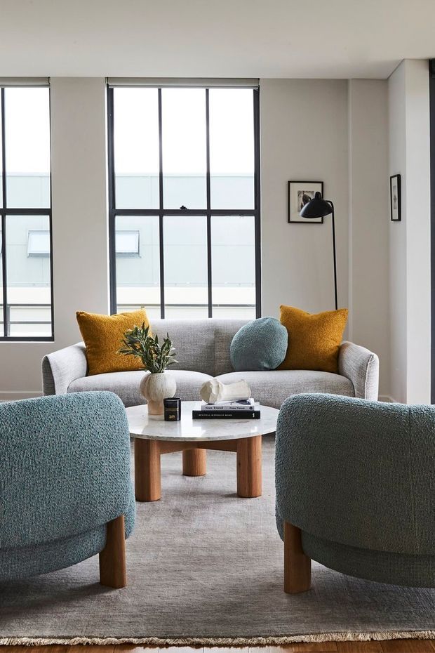 A bright, contemporary living room with a grey upholstered sofa and two rounded blue-grey armchairs arranged around a round timber coffee table. Mustard and blue cushions add colour, while a large neutral rug, framed artwork, black floor lamp, and tall black-framed windows create a calm, refined interior.