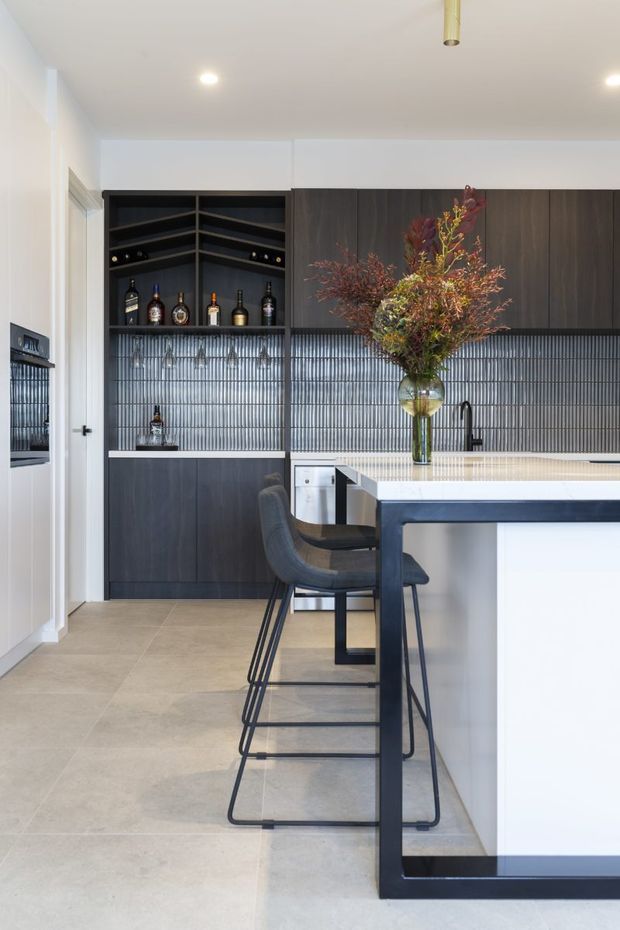 Contemporary kitchen featuring a large white island with a dark metal frame, charcoal bar stools, dark timber cabinetry, open bottle shelving, and a vertically ribbed dark splashback. Large-format pale flooring and white walls create a bright, refined interior, with a floral arrangement adding a natural accent.