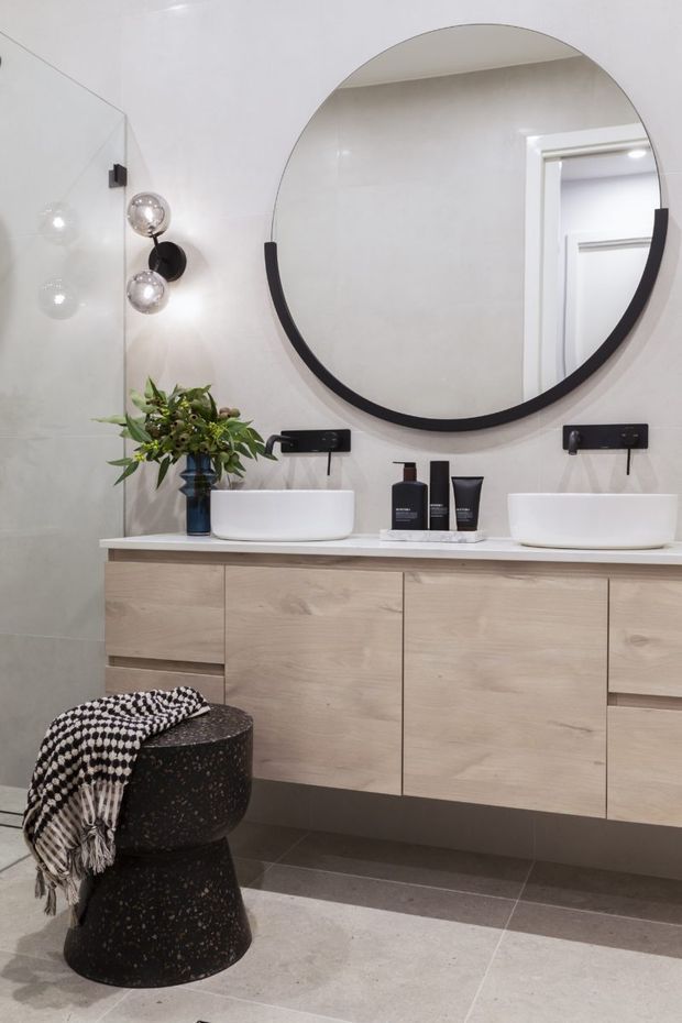 Contemporary double-vanity bathroom with a pale timber floating cabinet, white vessel basins, large round black-framed mirror, wall-mounted black tapware, and a light tiled interior. A dark terrazzo stool and botanical arrangement add contrast and texture.