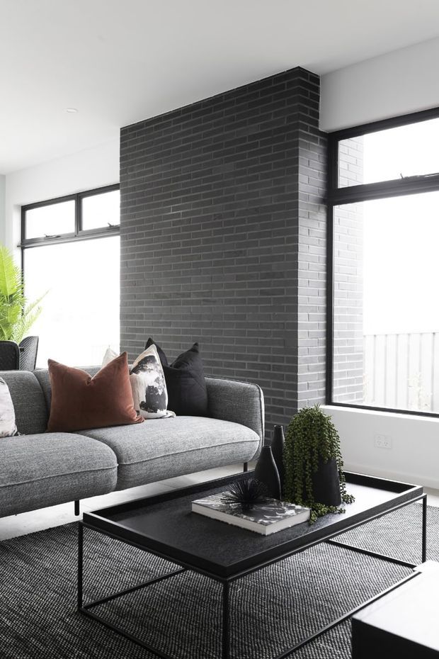 Contemporary living room with a large charcoal-painted brick feature wall, black-framed windows, a grey sectional sofa, and a minimalist black coffee table. Neutral furnishings are accented by rust and black cushions, sculptural dark vases, greenery, and a textured monochrome rug. The space is bright, refined, and high-contrast.