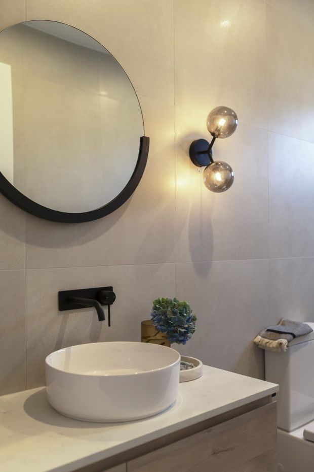 A contemporary bathroom vanity with a white vessel basin on a pale stone benchtop, light wood cabinetry, large round black-framed mirror, and black wall-mounted tapware. Beige tiled walls, twin spherical wall lights, and a toilet are visible.