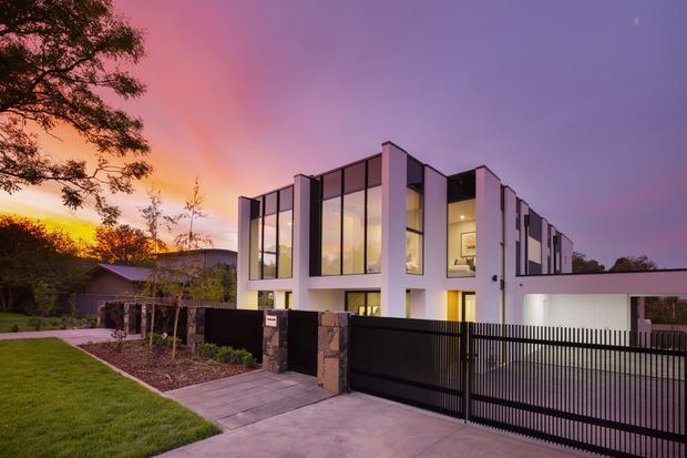A contemporary multi-storey residential house photographed at dusk, featuring a crisp white facade, extensive floor-to-ceiling glazing, strong vertical forms, and a flat roofline. A black metal slatted security gate and fence sit between stone-clad entry pillars, with a paved driveway and landscaped front yard. Warm interior lighting contrasts with the purple and orange sunset sky.