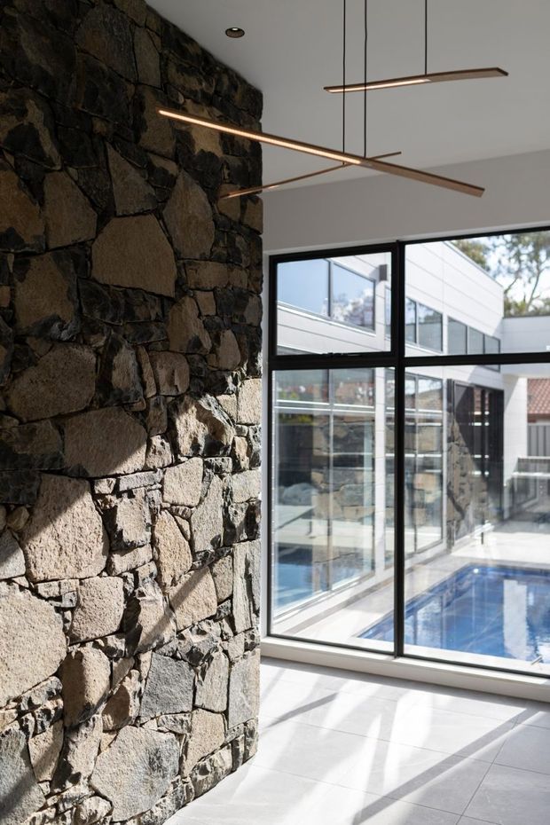 A bright contemporary interior featuring a rugged dark natural-stone feature wall, large black-framed floor-to-ceiling glazing, and a polished linear pendant light. A blue swimming pool and a modern white-clad extension are visible outside through the glazing. Pale large-format floor tiles and strong sunlight create a high-contrast, open atmosphere.