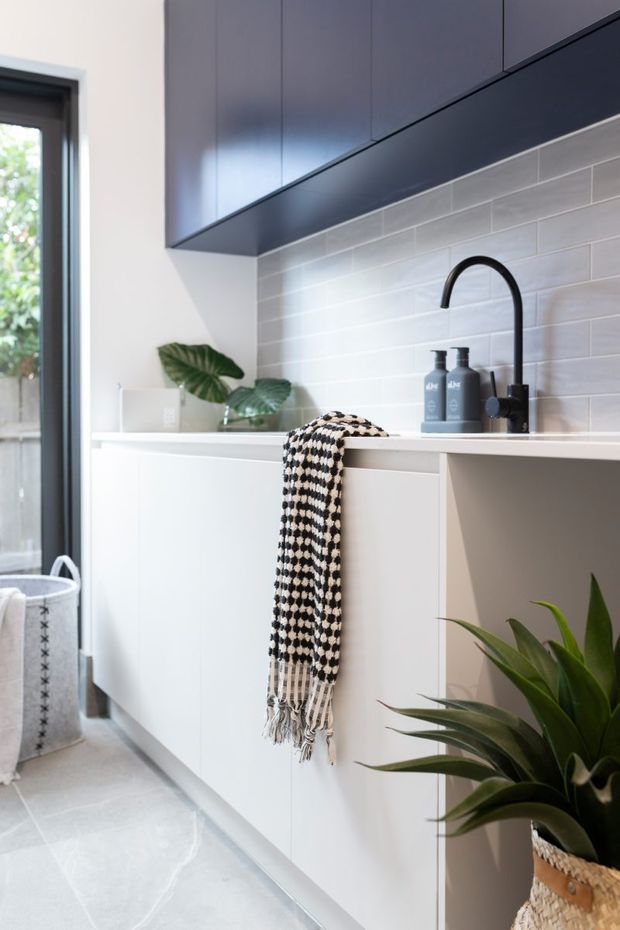 Bright contemporary laundry with extensive white built-in cabinetry, dark upper cabinets, a white subway-tile backsplash, black gooseneck tapware, and a pale tiled floor. A patterned textile hangs over the cabinetry, with indoor plants and a glazed exterior door adding natural detail.