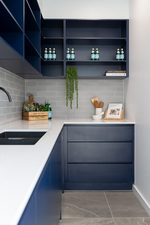 Compact contemporary kitchen with deep navy-blue cabinetry, open shelving, white benchtops, and a light grey subway-tile splashback. The L-shaped layout includes a dark undermount sink, matte drawer fronts, a trailing plant, and styled shelving with bottles and accessories. Large grey floor tiles and restrained styling create a clean, refined look.