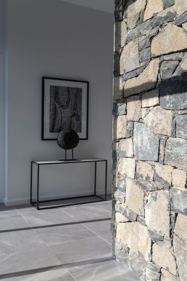 A minimalist interior entry or hallway with pale grey walls and large grey tiled flooring. A slim black metal console table supports a sculptural black object, with monochrome artwork above. A prominent rough stacked-stone structural or feature wall forms the right side of the frame, creating strong contrast with the restrained contemporary fit-out.