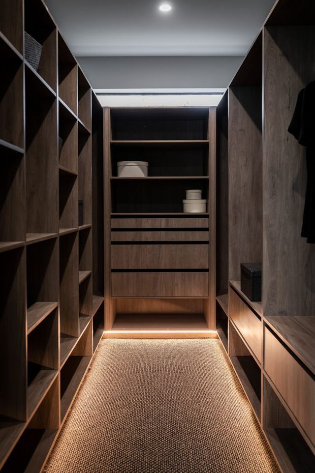 A narrow, highly organized walk-in wardrobe lined with dark timber built-in shelving, drawers, hanging storage and a central display unit. Warm integrated lighting illuminates the timber joinery and textured neutral runner, creating a refined, intimate dressing space.