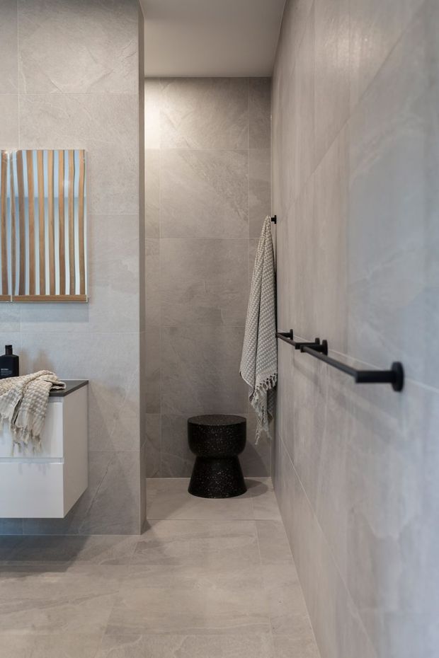 A contemporary bathroom with large-format light grey stone-look tiles covering the walls and floor. The space includes a floating white vanity, a black vessel basin, matte black towel rail, a textured towel, and a decorative timber-framed mirror with vertical slats. The compact room has a clean, refined, neutral palette with strong black accents.