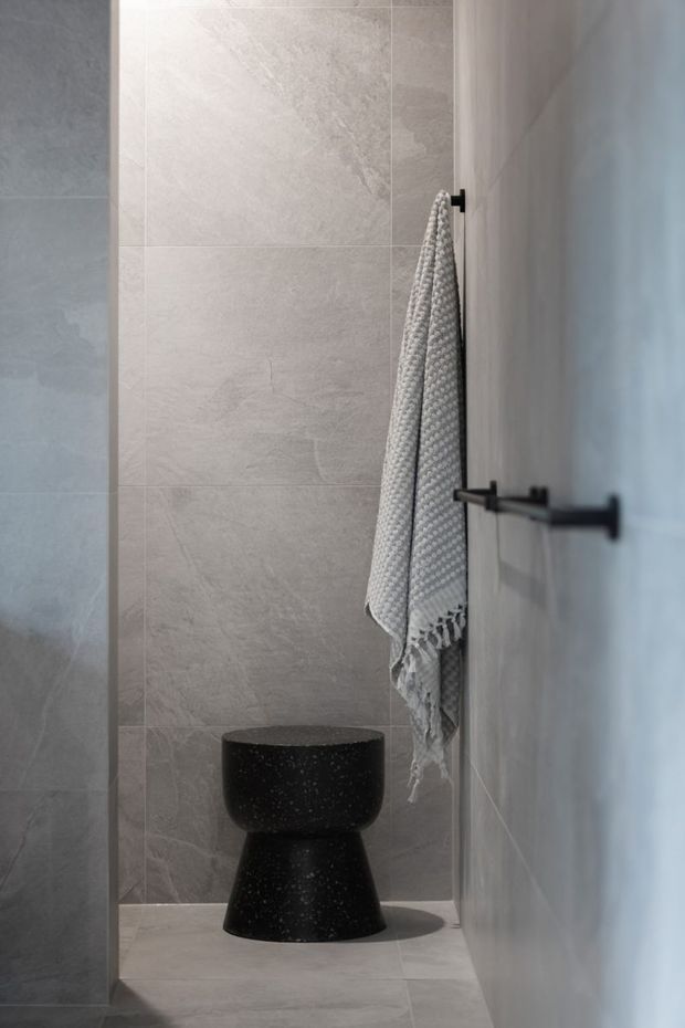 A narrow, minimalist bathroom featuring large light-grey stone-look wall and floor tiles, a sculptural matte-black freestanding toilet, and black wall-mounted towel hardware. A textured grey towel hangs beside the toilet, creating a restrained, monochromatic interior.