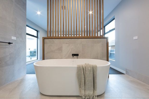 Bright contemporary bathroom featuring a freestanding oval bathtub positioned in front of a tiled feature wall with a wall-mounted tap. Vertical timber slats form a screen above the bath, while large windows, recessed ceiling lights, pale walls, and large-format light stone-look floor and wall tiles create an airy, refined space.