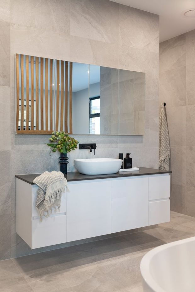 Contemporary bathroom featuring large-format light grey stone-look wall and floor tiles, a floating white vanity with dark benchtop, white vessel basin, wall-mounted black tapware, and a large mirror with vertical timber screening reflected behind it. A freestanding bath is partially visible in the foreground.