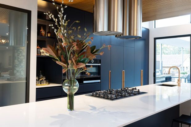 A sophisticated contemporary kitchen centered on a large white stone island with an integrated gas cooktop and a brass-toned tap. The kitchen features deep navy full-height cabinetry, black open shelving, built-in appliances, metallic rangehoods, and a warm slatted timber ceiling. Large glazed openings bring in abundant daylight and provide views outside. A vase of foliage sits on the island.