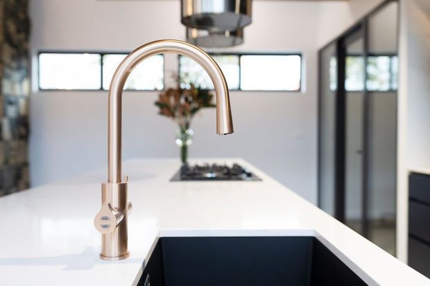 Close-up view across a bright contemporary kitchen island, featuring a tall brushed metallic gooseneck tap and a deep black undermount sink set into a white island benchtop. A cooktop, vase, horizontal clerestory windows and dark-framed glazing are visible in the background.