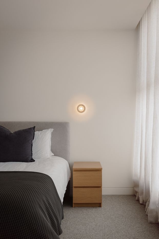 A calm, minimalist bedroom corner with a grey upholstered bedhead, white bedding, dark charcoal pillows and bedspread, a simple light timber three-drawer bedside table, pale neutral walls, grey carpet, and full-height sheer curtains. A small circular wall light provides a warm focal accent.