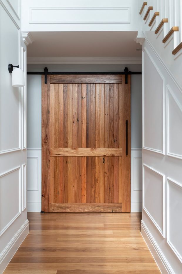 A refined residential hallway featuring a large rustic timber barn-style sliding door, warm timber flooring, white decorative wall panelling, and a staircase with exposed timber handrail brackets. A black wall sconce provides a contrasting accent.
