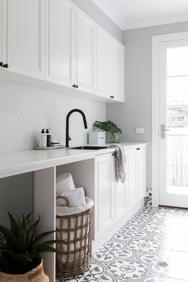 Bright, refined laundry room with white raised-panel cabinetry, a white countertop and integrated sink, black gooseneck tapware, patterned encaustic-style floor tiles, and a herringbone tiled backsplash. A woven laundry basket, folded textiles, and potted plants add warmth. The room opens to a glazed exterior door and is finished with light grey walls and decorative ceiling moulding.