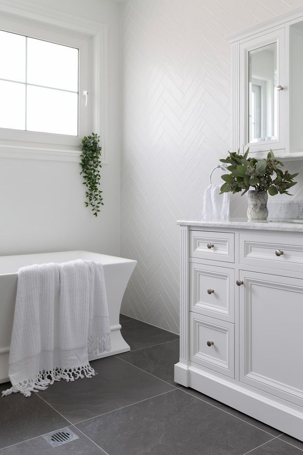 Bright, refined bathroom featuring a freestanding white bathtub, white shaker-style vanity cabinetry with brushed metal knobs, a pale marble-look benchtop, white herringbone wall tile, dark large-format floor tiles, and a mirrored wall cabinet. Natural greenery and white towels add soft accents.