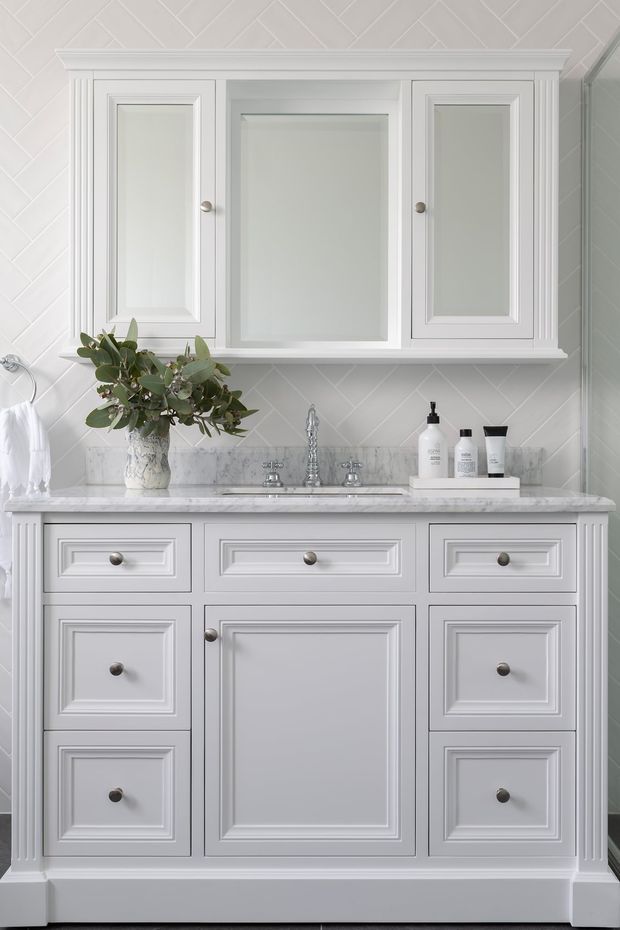 A bright, refined bathroom vanity wall featuring a large white painted timber vanity with paneled drawers and doors, a marble countertop, integrated double-basin fixtures, and a matching three-section mirrored wall cabinet. White diagonal tilework, decorative trim, and brushed metal hardware create a classic, polished appearance.
