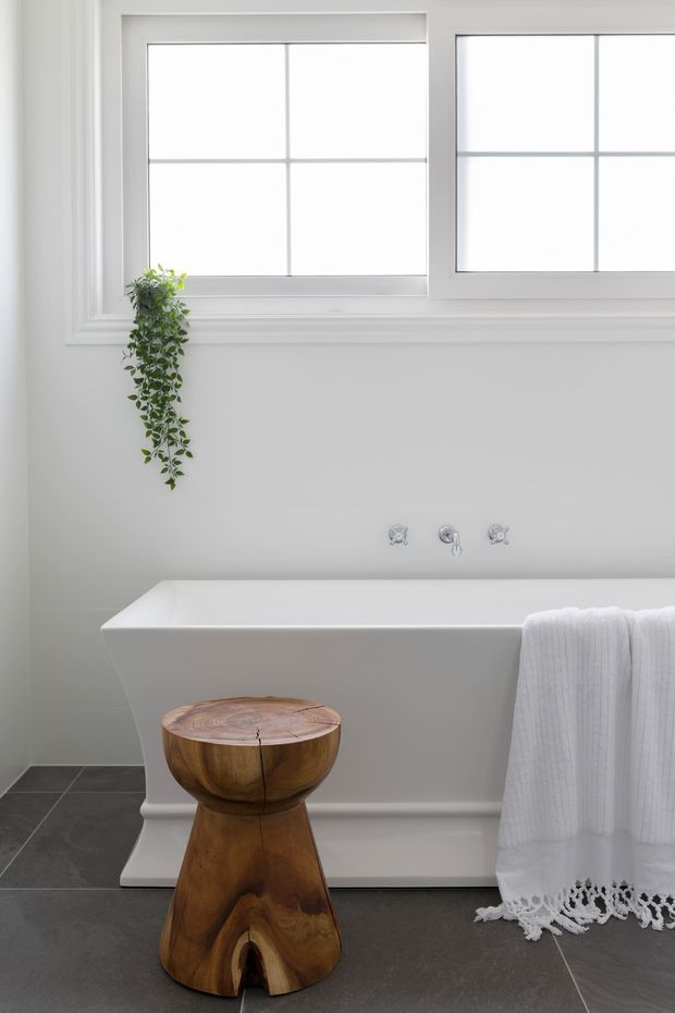 Bright, serene bathroom featuring a freestanding white bathtub beneath large white-framed divided windows. A small trailing plant adds greenery, while a sculptural timber stool and neatly draped white towels provide natural and textural accents. The room has pale walls, white moulded trim, and dark large-format floor tiles.
