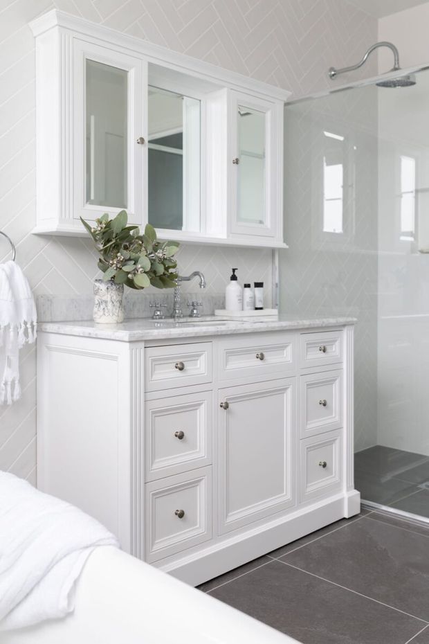 Bright, refined bathroom featuring a white paneled vanity with marble benchtop, mirrored wall cabinet, chrome tapware, white herringbone wall tiles, and large dark grey floor tiles. A frameless glass shower is visible to the right, while the edge of a white bathtub appears in the foreground.