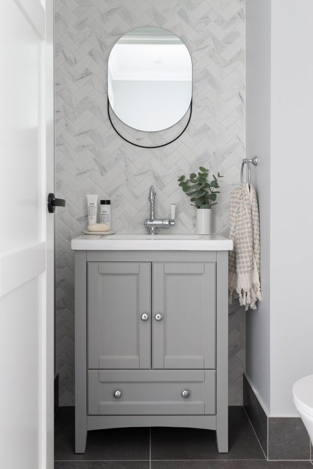 Compact, bright bathroom featuring a grey traditional-style vanity with paneled cabinetry, white benchtop, chrome wall-mounted tapware, dark floor tiles, and a white marble-look herringbone tiled feature wall. An arched black-framed mirror, folded hand towel, and small plant add decorative detail.
