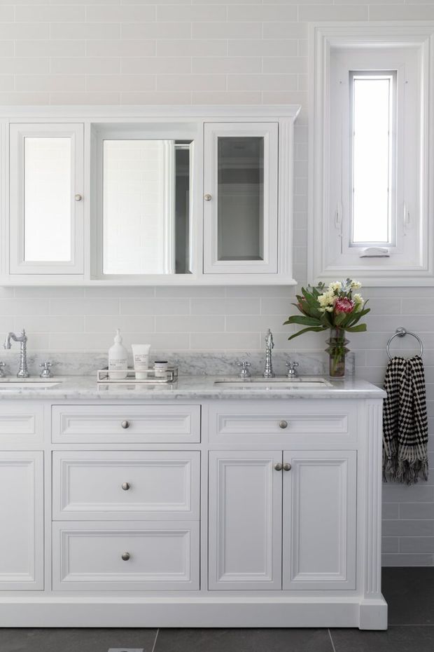 A bright, refined traditional bathroom vanity with white shaker-style cabinetry, framed mirrored wall cabinets, a marble benchtop, pale subway-tile walls, and polished chrome fixtures. A narrow white-framed window provides natural light, while a floral arrangement and patterned towel add small decorative accents.