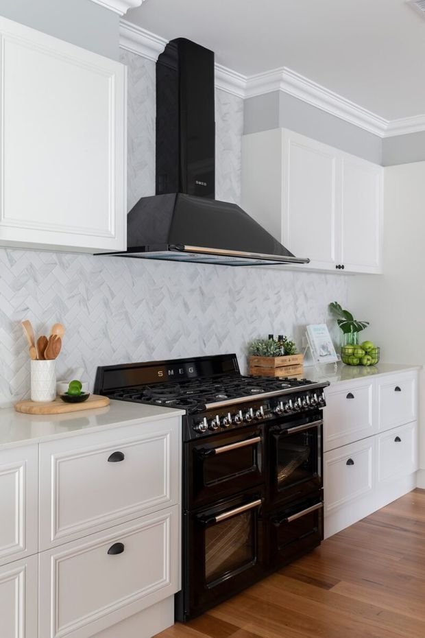 Bright, refined kitchen featuring white Shaker-style cabinetry, a large black range cooker and matching sculptural rangehood, pale herringbone backsplash, light stone benchtops, grey walls with decorative crown moulding, and warm timber flooring.