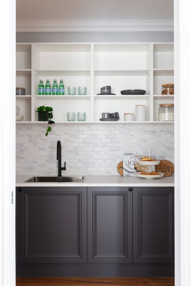 Compact, refined pantry or butler’s pantry with dark charcoal raised-panel base cabinetry, white engineered-looking benchtop, white elongated brick-pattern backsplash, open white shelving, and a black gooseneck tap over a small undermount sink. Glassware, bottled drinks, jars, dishes, and a trailing plant are neatly arranged on the shelves. The space has white trim and a warm timber floor, creating a crisp high-contrast finish.