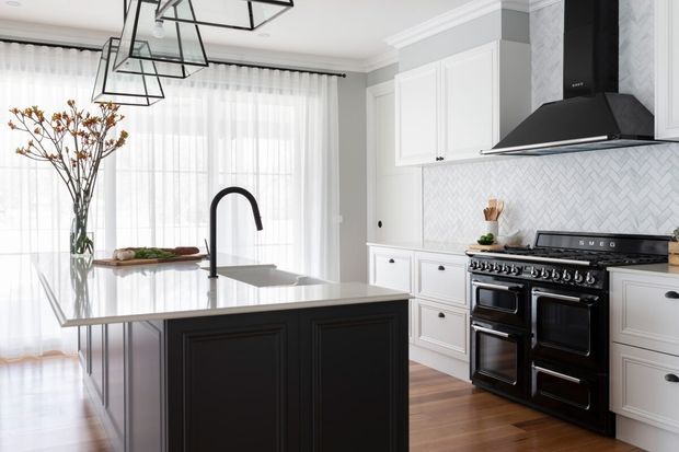 Bright, refined kitchen with a large black island, white perimeter cabinetry, a professional black range cooker and matching hood, white herringbone backsplash, timber flooring, and generous sheer-covered windows. Black geometric pendant lights and a sculptural black tap provide graphic contrast.