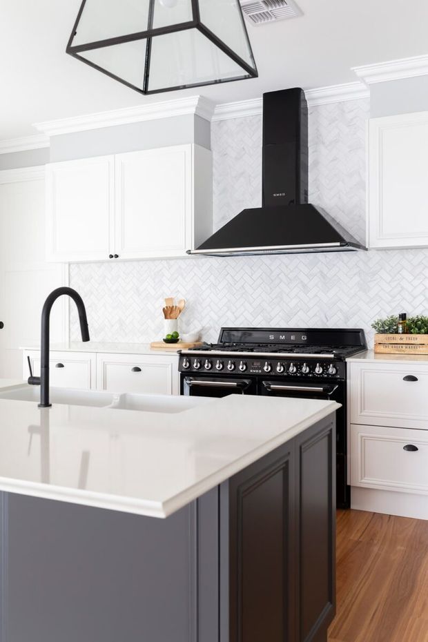 Bright, refined kitchen featuring white raised-panel cabinetry, a large dark island with a white stone benchtop, a black farmhouse-style range cooker and matching sculptural rangehood. The walls are finished with a white herringbone tile splashback, while timber flooring, black hardware, a matte black gooseneck tap and a geometric glass pendant add contrast. Decorative crown moulding gives the room a classic character.