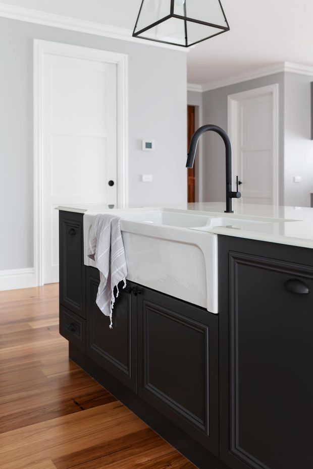 A bright, refined kitchen interior featuring a large black painted island with raised-panel cabinetry, a white farmhouse apron-front sink, white benchtop, and a tall matte-black gooseneck tap. The room has pale grey walls, white panelled doors and traditional trim, warm timber flooring, and a geometric black pendant light.