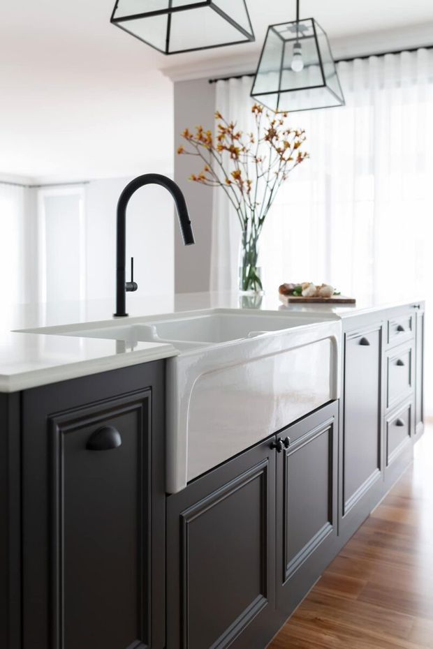 Bright, refined kitchen interior centered on a large island with a white farmhouse apron-front sink, matte black arched faucet, and dark raised-panel cabinetry with black hardware. White countertops, sheer curtains, geometric pendant lights, and warm timber flooring create a classic yet contemporary look.