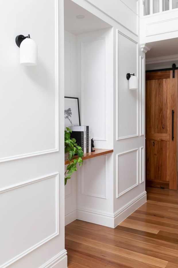 Bright, refined interior hallway featuring extensive white raised-panel wall moulding, matching white wall sconces, warm timber flooring, a recessed display nook with a timber shelf and greenery, and a substantial natural timber sliding barn door with black hardware. The space has a classic architectural character with contemporary restraint.