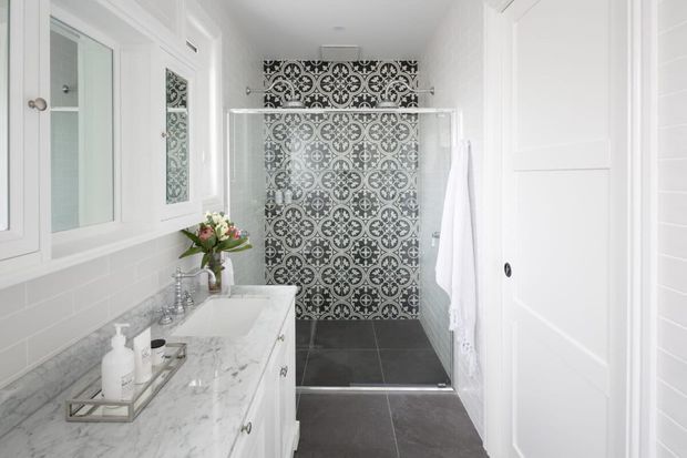 Bright white bathroom with a marble vanity and integrated basin, chrome fixtures, white subway-tile walls, dark grey floor tiles, and a walk-in shower featuring an ornate black-and-white patterned tile feature wall. White cabinetry, shutter-style doors, and a mirrored wall cabinet complete the refined traditional interior.