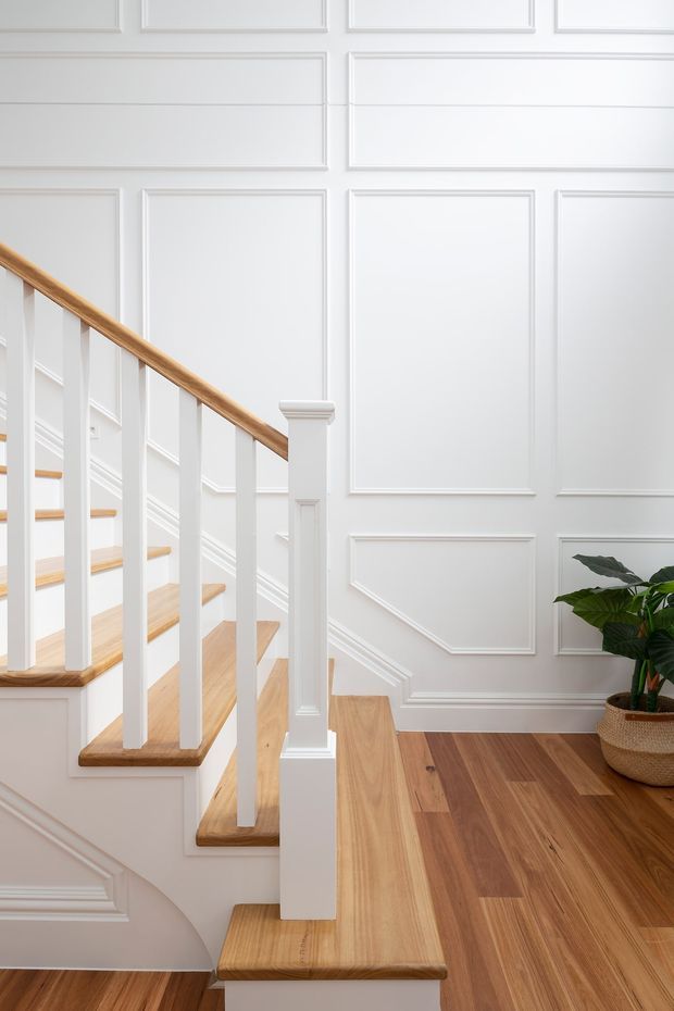 Bright, refined stairway interior featuring white raised-panel wall moulding, a white staircase with slim square balusters, warm timber stair treads and handrail, and wide-plank timber flooring. A potted plant sits at the base of the stairs.