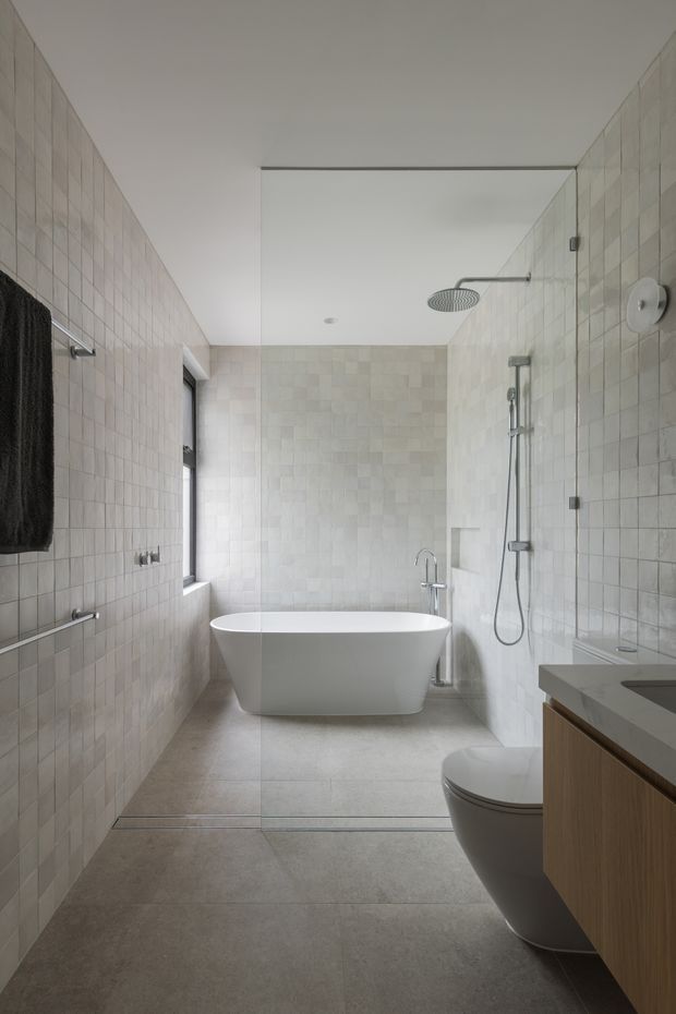 A bright, refined contemporary bathroom with full-height pale square wall tiles, large-format grey floor tiles, a freestanding white bathtub, frameless glass shower screen, wall-mounted shower fixtures, and a timber vanity with a grey stone benchtop. A wall-hung toilet, towel rails, and narrow window complete the clean, minimalist composition.