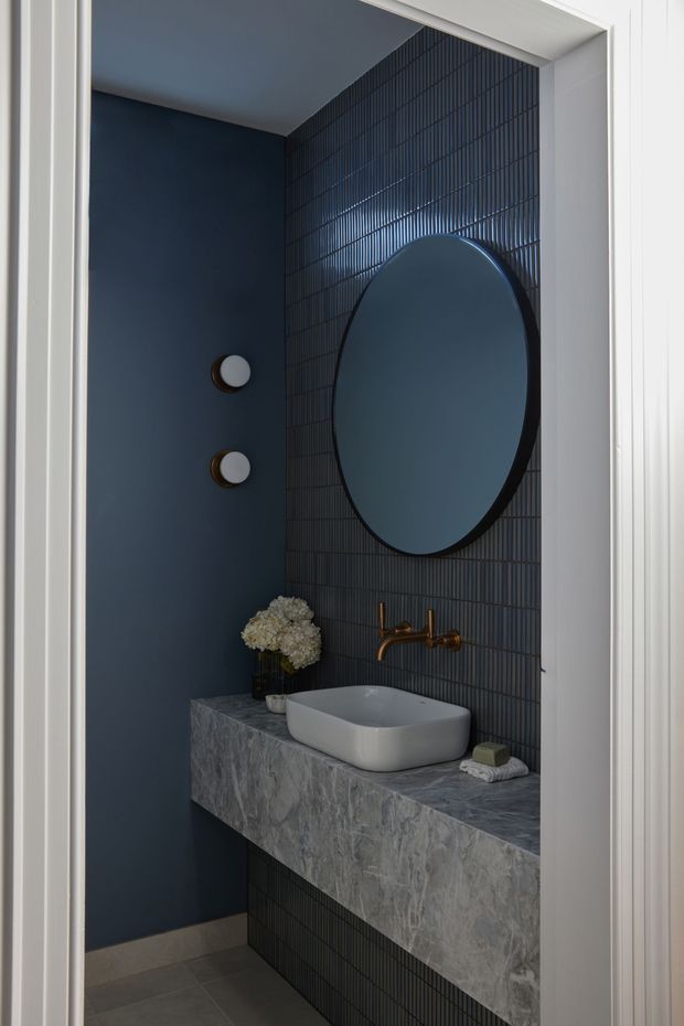 A compact, moody bathroom viewed through a doorway, featuring a floating grey stone vanity with a white vessel basin, bronze wall-mounted tapware, dark blue painted walls, and a dark vertically ribbed tile feature wall. A large oval mirror and two round wall lights add strong graphic elements.